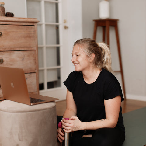 En kvinna sittandes på en yoga matta, med en vattenflaska i handen och en dator framför sig som sitter och pratar.