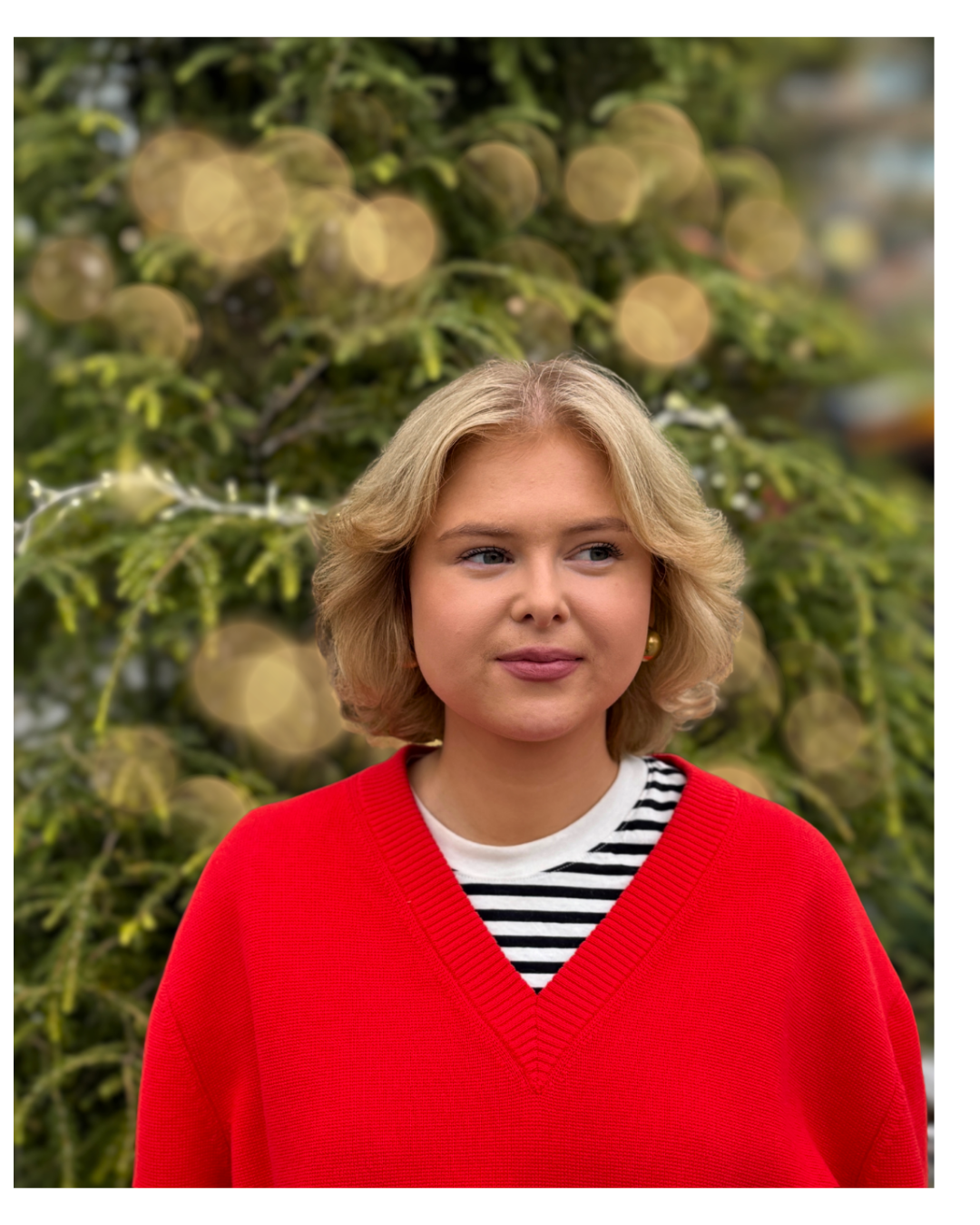 En tjej som ler iklädd en röd tröja som står framför en gran men en ljusslinga som hänger över grenarna