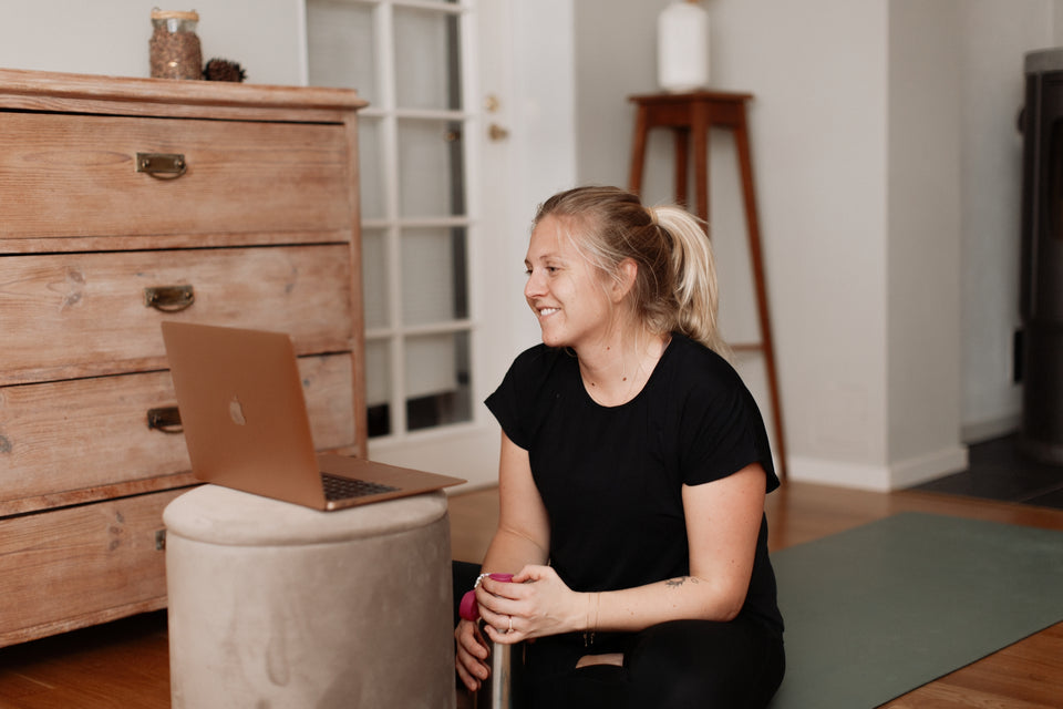 En kvinna sittandes på en yoga matta, med en vattenflaska i handen och en dator framför sig som sitter och pratar.